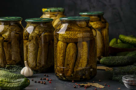 Different pickled cucumbers with zucchini, onions, garlic and grape leaves in jars on a dark background, horizontal photoの写真素材