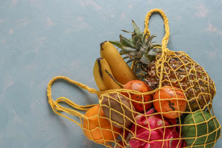 Zero waste concept, mesh textile bag with fresh tropical fruits: mango, pineapple, dragon, kiwi, banana and passion fruit on light blue background, horizontal orientationの写真素材
