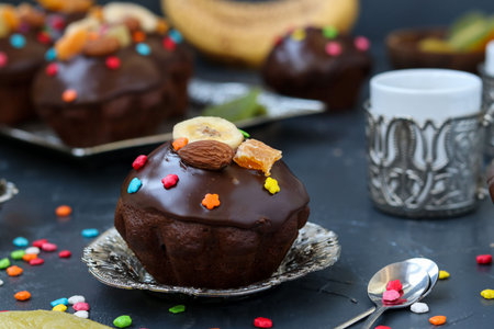 Banana-chocolate muffins, covered with icing and confectionery confetti, are located on a dark...