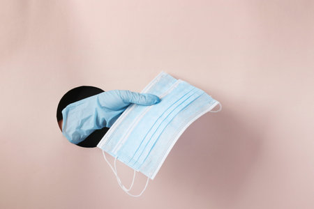 Female hand in a glove holds two protective medical masks through a round hole in pink paper, copy...