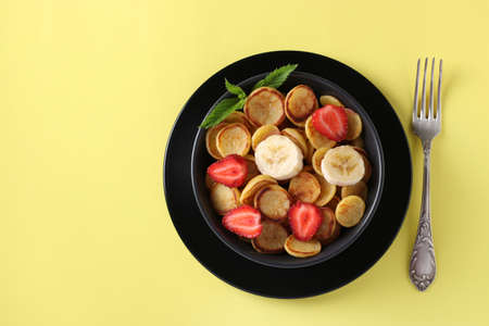Trendy tiny pancakes for breakfast with strawberry and banana in dark bowl on yellow background. Top view. Space for textの写真素材