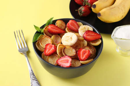Trendy tiny pancakes for breakfast with strawberry, banana and sour cream in dark bowl on yellow background. Closeupの写真素材