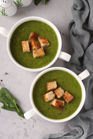 Green spinach puree soup served with croutons in two white bowls on a gray concrete background,...