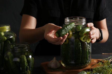 Marinated cucumbers with garlic, pepper and dill in a glass jars on dark background. Woman fills a jar with cucumbers.の写真素材