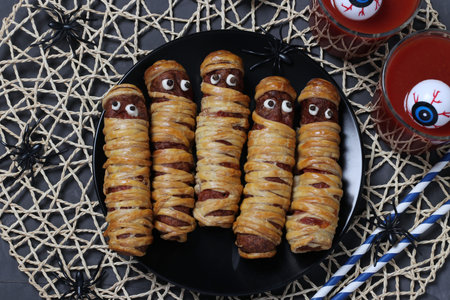 Spooky sausage mummies and tomato juice for Halloween party on dark plate, Closeup, Flat lay.