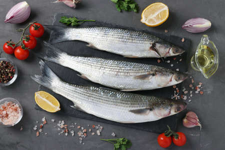 Fresh ready to cook raw mullets with ingredients and seasonings like parsley, lemon, cherry tomatoes, red onion and olive oil on dark background. Top view.の写真素材