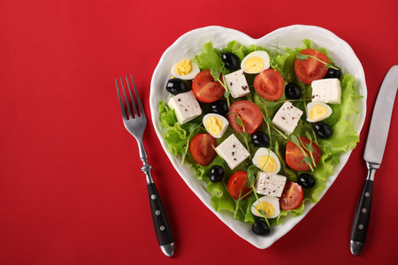 Healthy salad with cherry tomatoes, feta, quail eggs, black olives and microgreens peas on a heart-shaped plate on a red background. View from aboveの写真素材