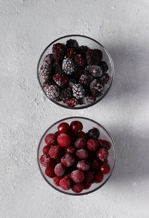 Frozen cherries and blackberries in two glass transparent bowls on gray concrete background. View...