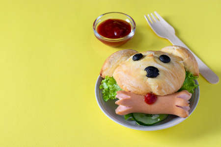 Children's sandwich in the shape of a dog, consisting of a bun, salad and sausage, on a yellow background with a place for textの写真素材