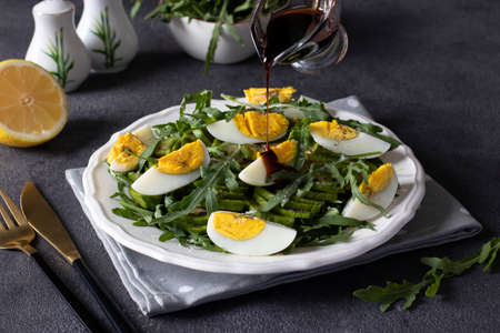 Healthy salad with avocado, arugula and eggs on a white plate on a dark background. The chef pours balsamic sauce over the salad.の写真素材