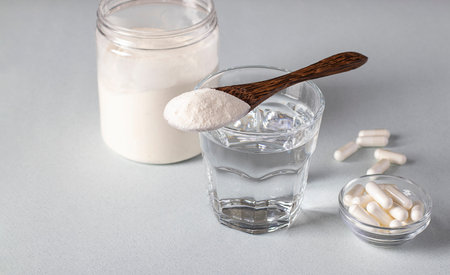 Collagen powder in spoon on glass of water, collagen capsules and jar of powder on a light...