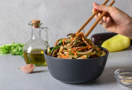 Korean style eggplant salad with sesame seeds, onions and carrots in a dark bowl on a light gray background.の写真素材