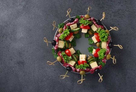 Festive canapes with sausage, cucumbers, tomatoes, olives and cheese, served on a plate as a Christmas wreath, on a dark background. Top viewの写真素材