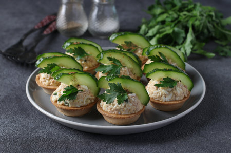 Tartlets with cod liver, egg, garnished with cucumber and parsley on plate on dark gray background
