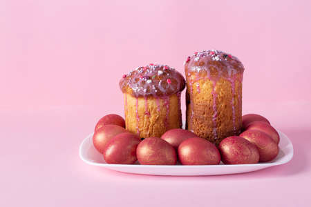 Two Easter cakes and colored eggs on white plate on pastel pink backgroundの写真素材