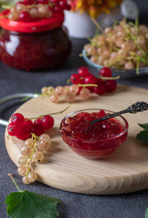 Homemade red and white currant jam in transparent bowl on wooden board, as well as jar of jam in...