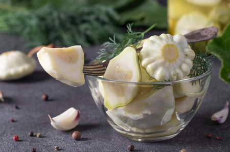 Pickled patissons with garlic in bowl and glass jar on dark gray table with fresh ingredients,...