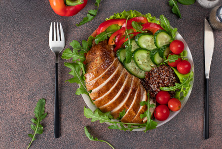 Salad with baked chicken breast, quinoa, cherry tomatoes, cucumbers, bell pepper, lettuce leaves and arugula on brown tableの写真素材