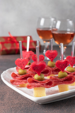 Canape for Valentines Day with hearts from cheese, olives and sausage on white plate shaped-heart and two glass rose wine on gray backgroundの写真素材