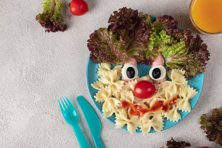 Fun Food for kids - smiling face from pasta, sausages, egg, cherry tomato and lettuce leaves on blue plate. Creative healthy breakfast for childrenの写真素材