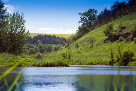 landscape consisting of a green grass on a foreground and river of the middle and green grassy hills with trees on the other side and forest with valley and blue sky with white clouds at distanceの写真素材