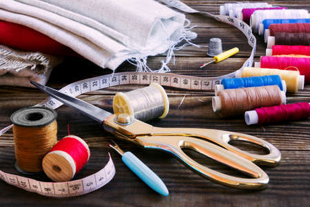A collection of sewing tools and different textile fabric and multi-coloured threads on a wooden brown backgroundの写真素材