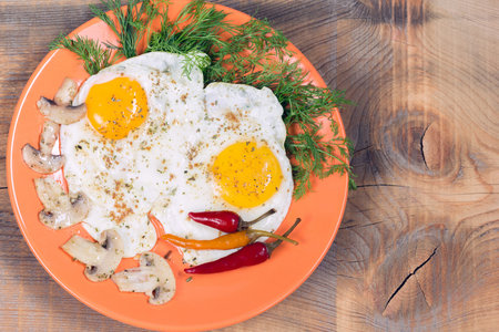 Fried two eggs with champignons, dill and red chili pepper on an orange plate on a wooden background
