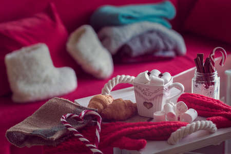 Warm and cozy winter still life, consist of, cup of coffee, marshmallow, croissants, lollipop on the foreground and fur shoes with  sweaters in the home interior living roomの写真素材