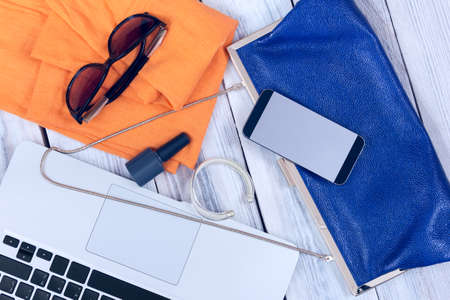 blue handbag and orange scarf with laptop and women's accessories on a white wooden backgroundの写真素材