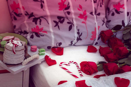 Coffee and cookie on a salver and red roses with his scattered petals on a bed with two pillows and heart of striped lollipopsの写真素材