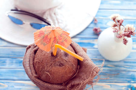 Ready to travel. Coconut with straw is wrapped in burlap on a blue background with white hat and blue sunglassesの写真素材