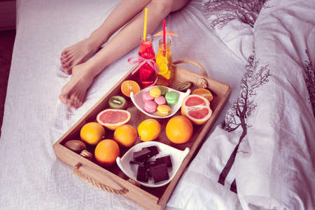 breakfast from different fruits and beverage to bed and female legs protruding from under the blanketの写真素材