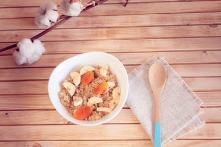 Quinoa porridge with banana and dried apricots on a wooden background with a sprig of cotton and...
