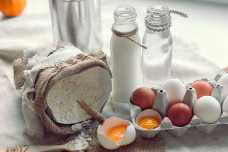 flour with spoon in a sack and broken eggs with milk and water on a backgroundの写真素材
