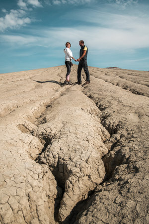 Young couple holding hands while staying on cracked dry soil, picture taken in the mud volcanoes park in Romaniaの写真素材