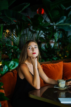 Beautiful young woman siting on soft sofa at table in cafe. Thought Girl looking into distance. Green plants in background. Hot drink cup on table. Young woman wearing black dress on thin strapsの写真素材