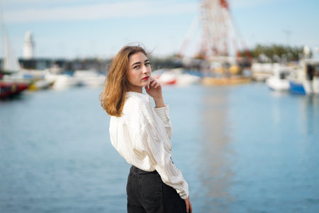 Beautiful girl standing near sea. Seaport or berth in background. Young woman Wearing casual Outfit...