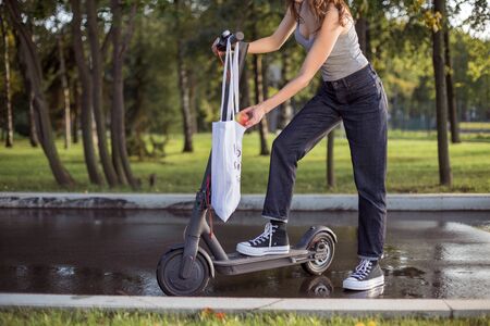 A brunette girl is standing next to electric scooter in the park and takes from a bagの写真素材