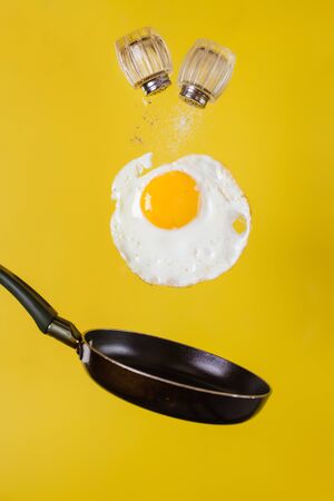 Breakfast time. A fried egg and a frying pan with salt shakers from which salt and pepper are strewed levitate in the air on a yellow backgroundの写真素材
