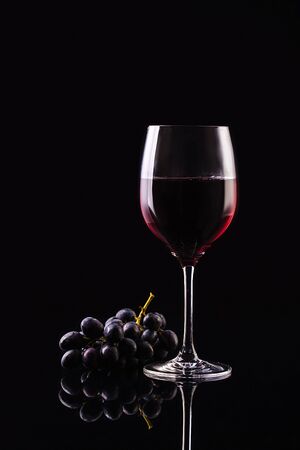 A a glass of wine on a black background with grapes are on a glossy surface. Aromatic wine. Strict styleの写真素材