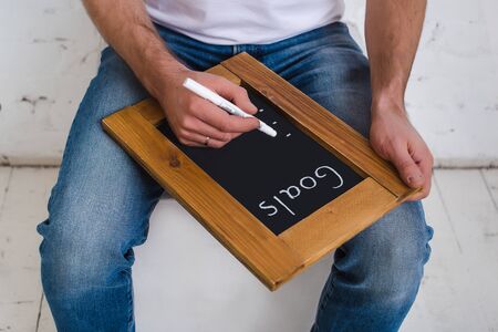 A man in a white T-shirt and blue jeans sits on a cube and is holding a white marker and a...