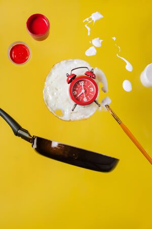 Breakfast time. Fried egg and frying pan with a red watch, paint jars and spray paints and a brush paint levitate in the air on a yellow backgroundの写真素材
