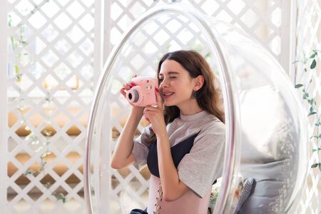 The girl sits in a transparent chair and takes a photo on the instant camera