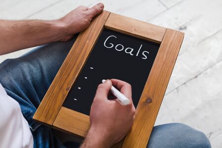 A man in a white T-shirt and blue jeans is holding a white marker and a whiteboard in a wooden frame for writing new goals. Close-up without a faceの写真素材