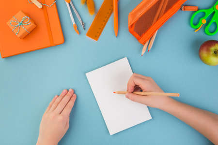 Child hand writes on white peace of paper school or office workspace with orange supplies on blue...