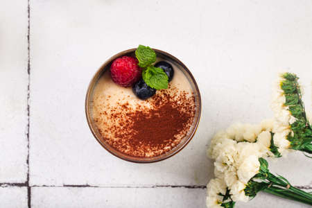 tiramisu dessert in glass with berries on white tile background with flowers