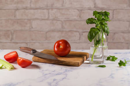 Womans hands in white apron black gloves cutting tomato on green board, behind fresh vegetables.
