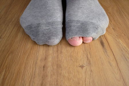 Close up of female feet wearing gray socks on wooden floor background. Wearing worn out socks with a hole and visible finger and toe sticking out. Poverty due to inflation.の写真素材