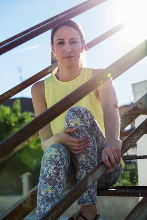 Relaxed and thoughtful young woman on the stairs from the rooftop of his house looking at camera through the railing  Attractive young woman sitting on staircase looking at camera waiting and relaxingの写真素材