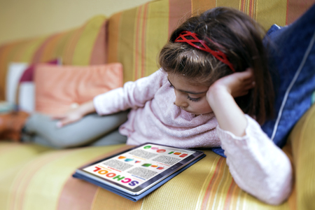 girl student reading relaxed in her tablet computer the school work / education concept. Little girl lying on the sofa at home studying home work on the tabletの写真素材
